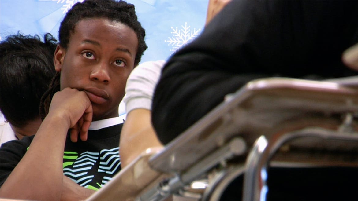 Close-up of teenage, African-American male sitting in a desk and looking towards the front of the classroom, looking mildly bored.