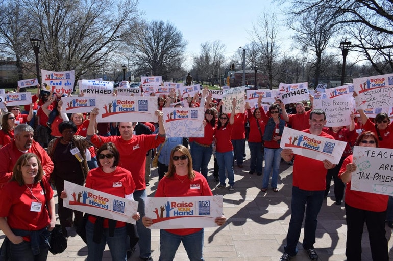 Photo of group of people outside in red shirts holding signs.