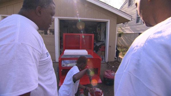 Two men watch as another paints a hot dog cart in front of a garage.