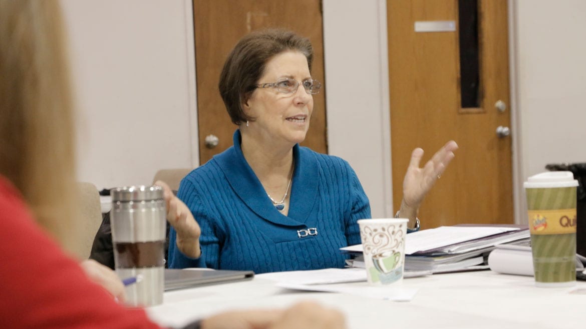 a woman in a blue sweater gestures with her hands as she speaks in a meeting