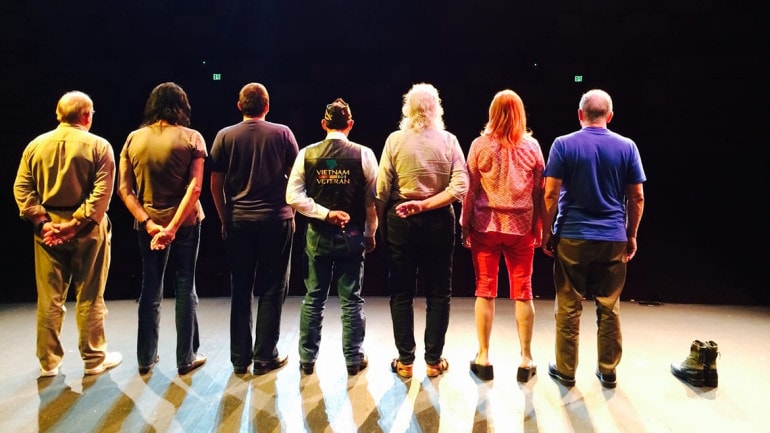 Photo of the backs of seven people standing on a stage.