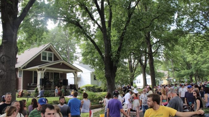 A large crowd moves down the street, taking in live music from a nearby porch.