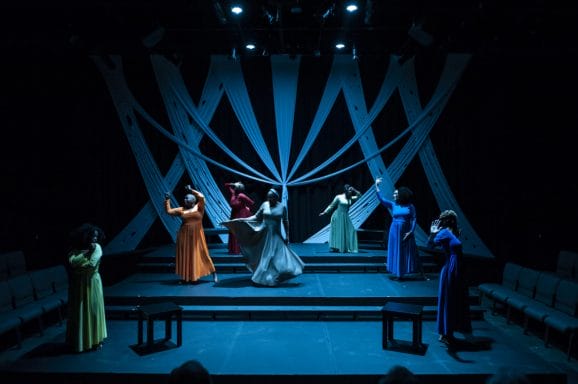 Seven women perform on a dim blue stage. 