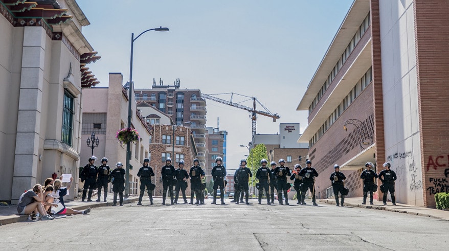 A row of police line up in the middle of the street, flanked by demonstrators on the left and right.