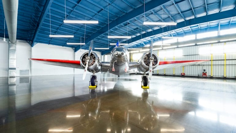 A silver plane with red wings and twin engines sits in an otherwise empty hangar
