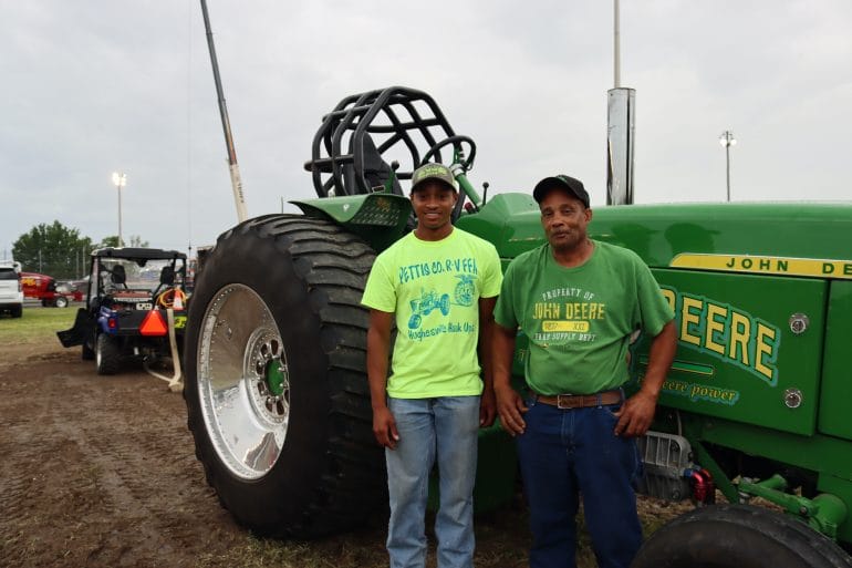Two men wearing green shirts and hats stand in front of a John Deere green tractor.