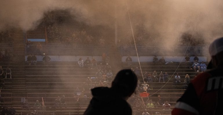 Two silhouettes look out at the grandstands as exhaust smoke settles in the air.
