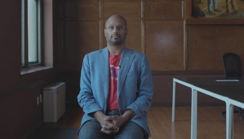 Man with glasses sits in a wood paneled room. He is wearing a blue blazer and red tshirt.