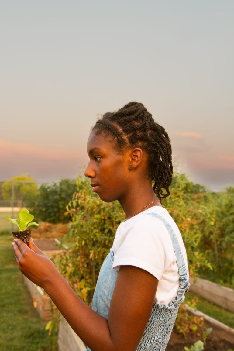 A young Black girl, Zara Ba, is standing between raised beds against the Kansas City sky as the sun goes down, while holding onto her lettuce. Photo by Ji Stribling for Flatland.