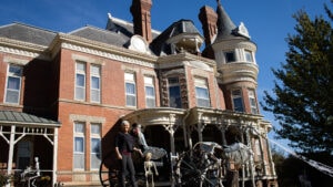 Woman stands next to old carriage with skeleton horses. Behind her is a victorian style mansion with brick facade and white trimming.