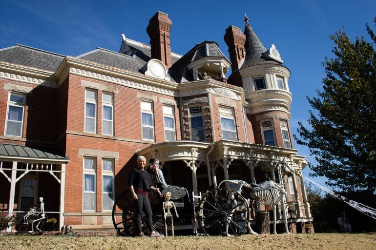 Woman stands next to old carriage with skeleton horses. Behind her is a victorian style mansion with brick facade and white trimming.