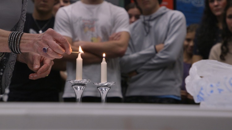 A elderly woman holds a match over a white candlestick. She's wearing a large amethyst ring. High schoolers watch in the background.