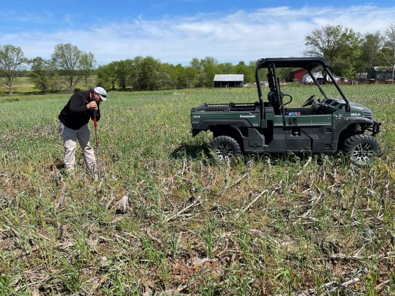 A man stands in a field that has dead corn stalks and a young cover crop. He is digging for a soil sample. Next to him is a green four wheeler.