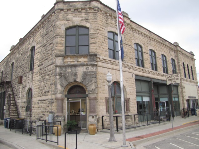 A limestone building on the corner of a street with an American flag on a pole next to it. 