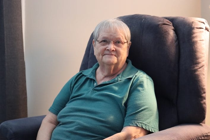 An elderly woman with short white hair and glasses smiles from her recliner.