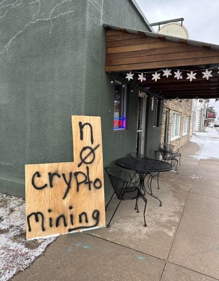 piece of scrap wood leans up against a building. There are tables and chairs outside of the restaurant and an "open" sign in the window. The scrapwood is spraypainted with "No Crypto Mining" 