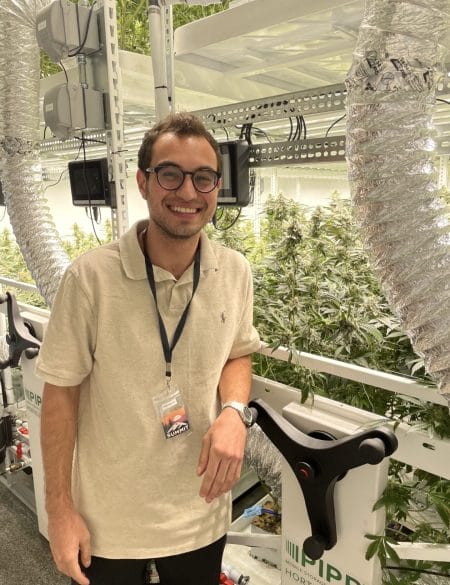 A man in glasses stands in a sterile looking facility. Behind him are rows of cannabis plants.