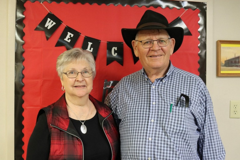 and older couple stand in front of a red bulletin board that reads "welcome"