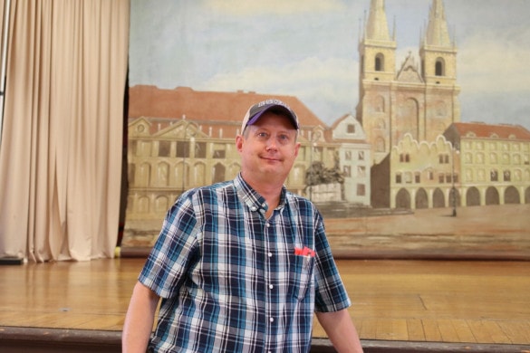 a man in a ball cap and plaid shirt stands in front of a painted backdrop.