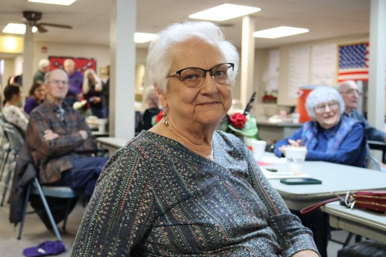 A woman with glasses smiles. Other folks look past her in the background but aren't in focus.