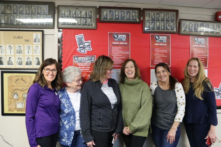A group of ladies of varying ages gather in front of a red bulletin board with information about the Rock-A-Thon