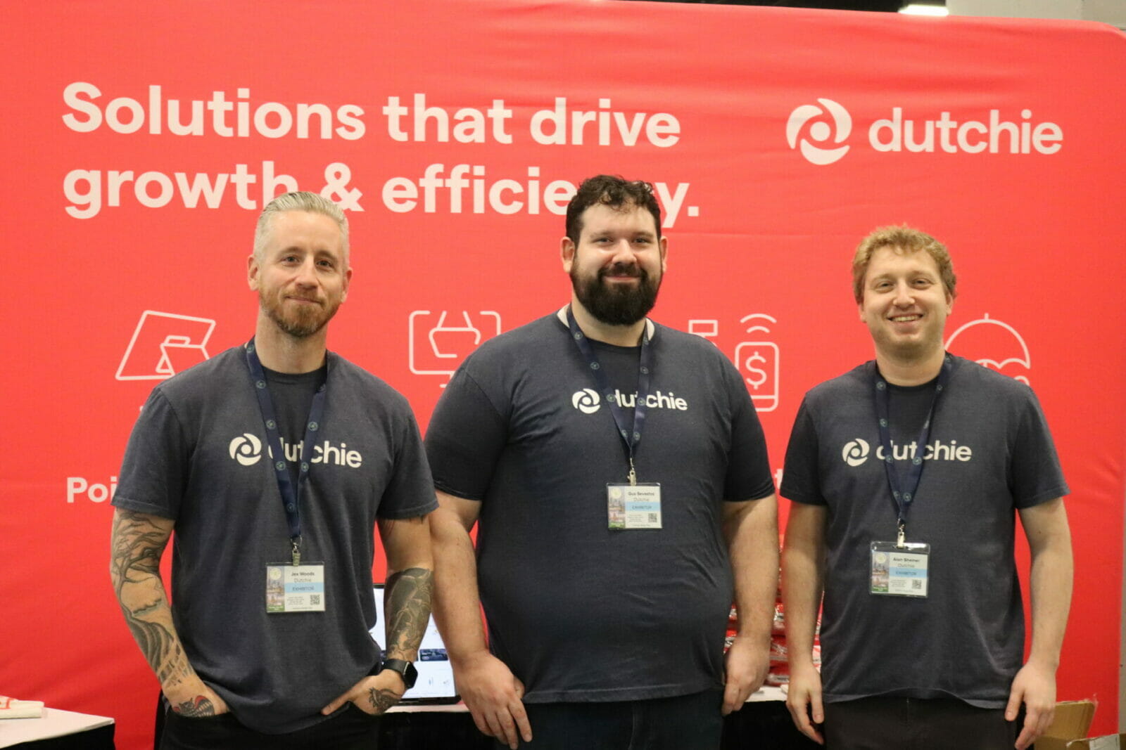 Three men stand in front of a red banner with white marking and drawings. Each of them wear a lanyard and a gray t-shirt which reads "dutchie."