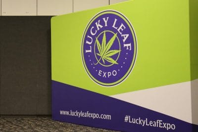 A green and blue sign in the corner of a room reads "Lucky Leaf Expo" with a cannabis leaf in the middle.