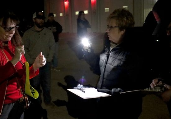 A woman in a black coat holds up a cellphone with the flashlight on. Her other hand has a clipboard and pen. Next to her someone signs a petition and others mingle around.