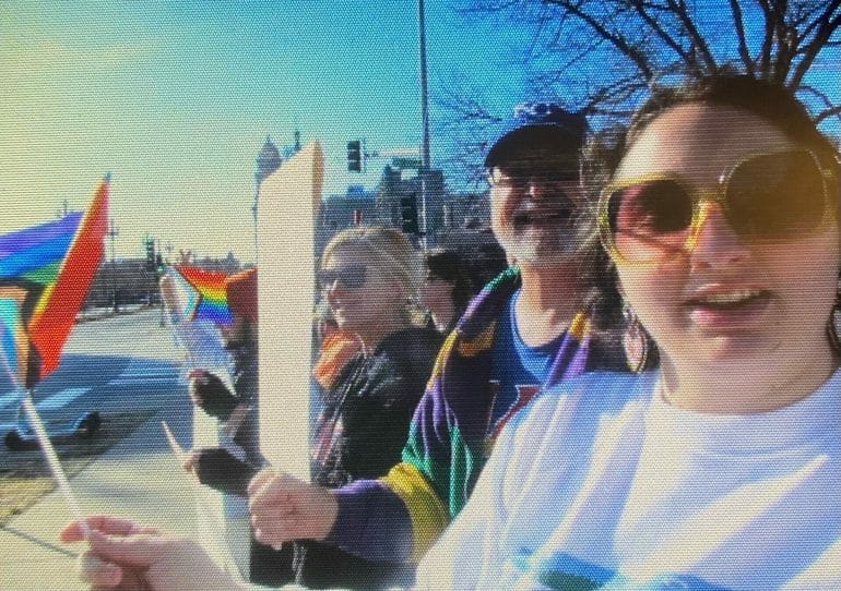 An attendee snaps a selfie with fellow protest attendees, all holding mini pride flags.