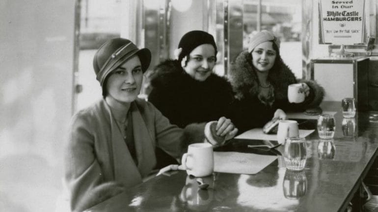 Three young women enjoying coffee and hamburgers at the counter of an unidentified White Castle restaurant, ca. 1930-1939.
