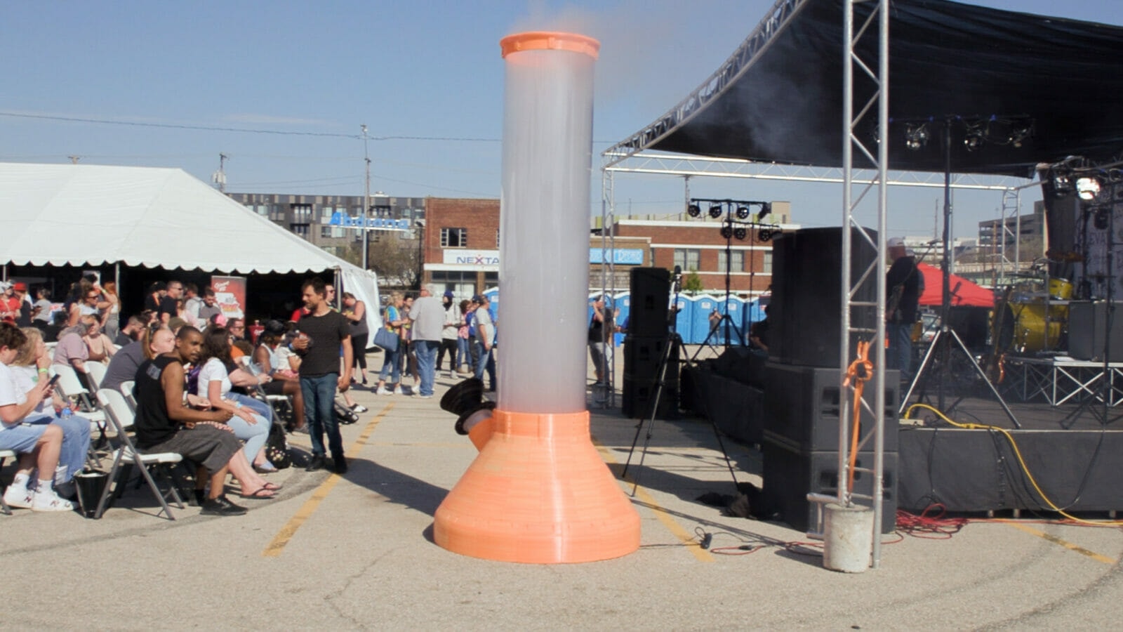 A giant, orange bong is filled with smoke. It's taller than the stage next to it. A crowd of people sit to the left.
