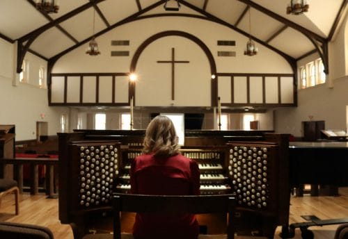 From behind, a woman sits at an organ with four rows of keys and many white knobs on the sides. In front of her is a sanctuary with white walls, stained glass and dark wood trimmings. A cross hangs in the center.