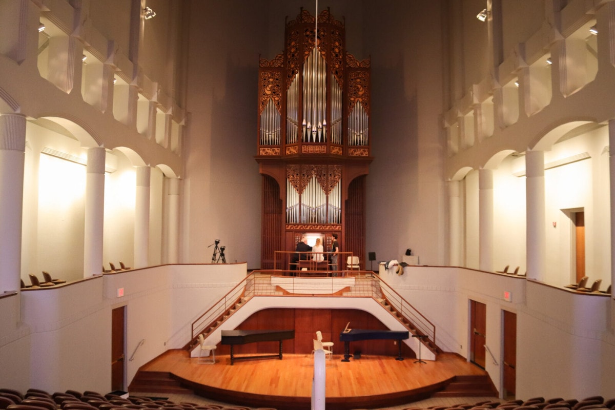 In a white concert hall, at the front of the hall sets a massive, ornate organ with three people at its console.