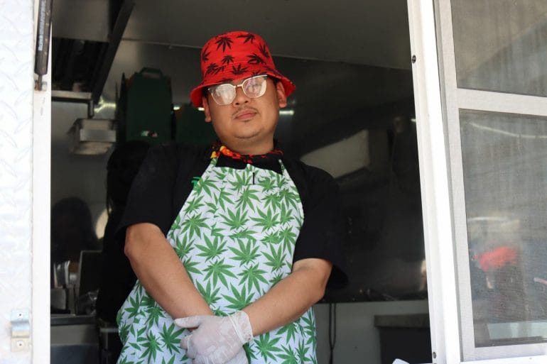 a man stands in the window of a food truck with a cannabis leaf bucket hat and apron on.