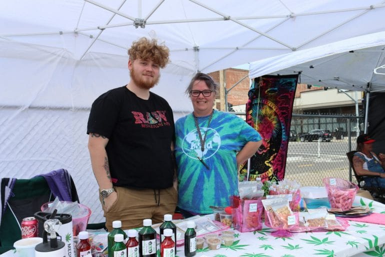 An older woman stands next to a younger man. In front of them is a table with a cannabis leaf table cloth and packaged, baked edibles.