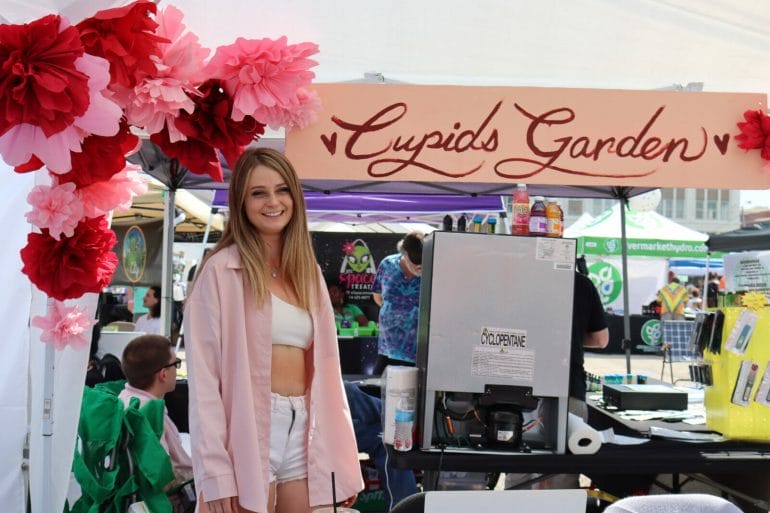 A girl in a pink shirt stands next to a sign that reads "Cupid's Garden" next to her are pink and red pom poms hung from the top of a portable tent.