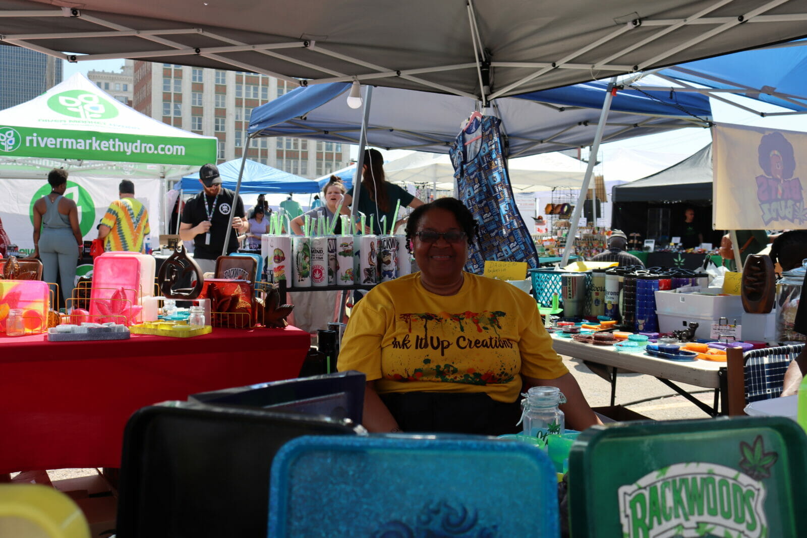 A woman in a yellow shirt that reads "Ink It Up Creations" around her are colorful rolling trays.