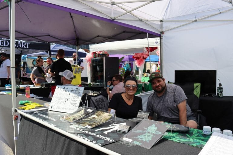 Two people sit under a tent behind a table that displays photos of cannabis flowers.