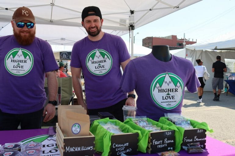 Two men in hats and purple shirts that read "higher love" stand behind a table of edibles. Signs on the edibles read "marshmallows, chocolate chip cookies, fruity pebble treats, fudge brownies"