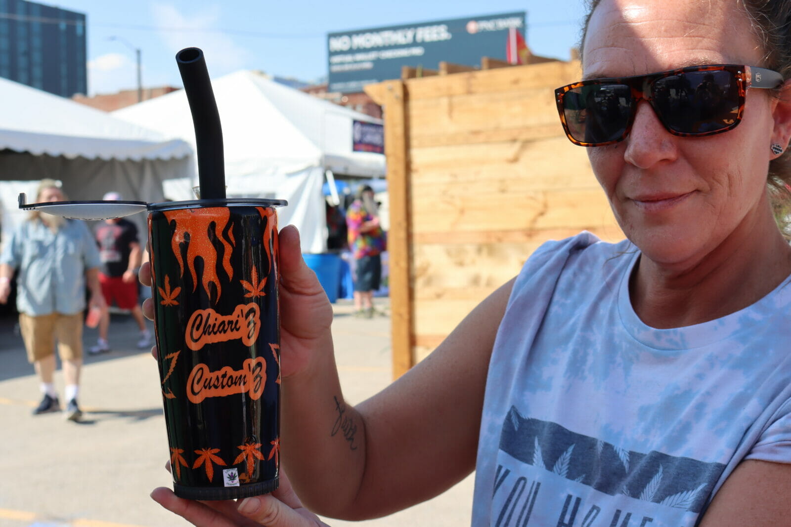 A woman holds up a cup decorated with Harley Davidson flames and cannabis leaves. In the center it reads "Chiari'z Custom'z" the cup has a straw and a bowl attached to it.