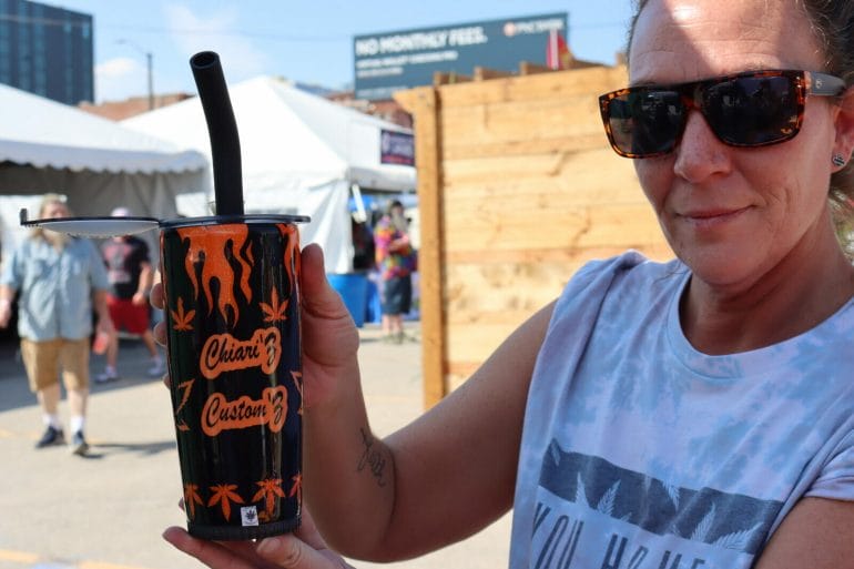 A woman holds up a cup decorated with Harley Davidson flames and cannabis leaves. In the center it reads "Chiari'z Custom'z" the cup has a straw and a bowl attached to it.