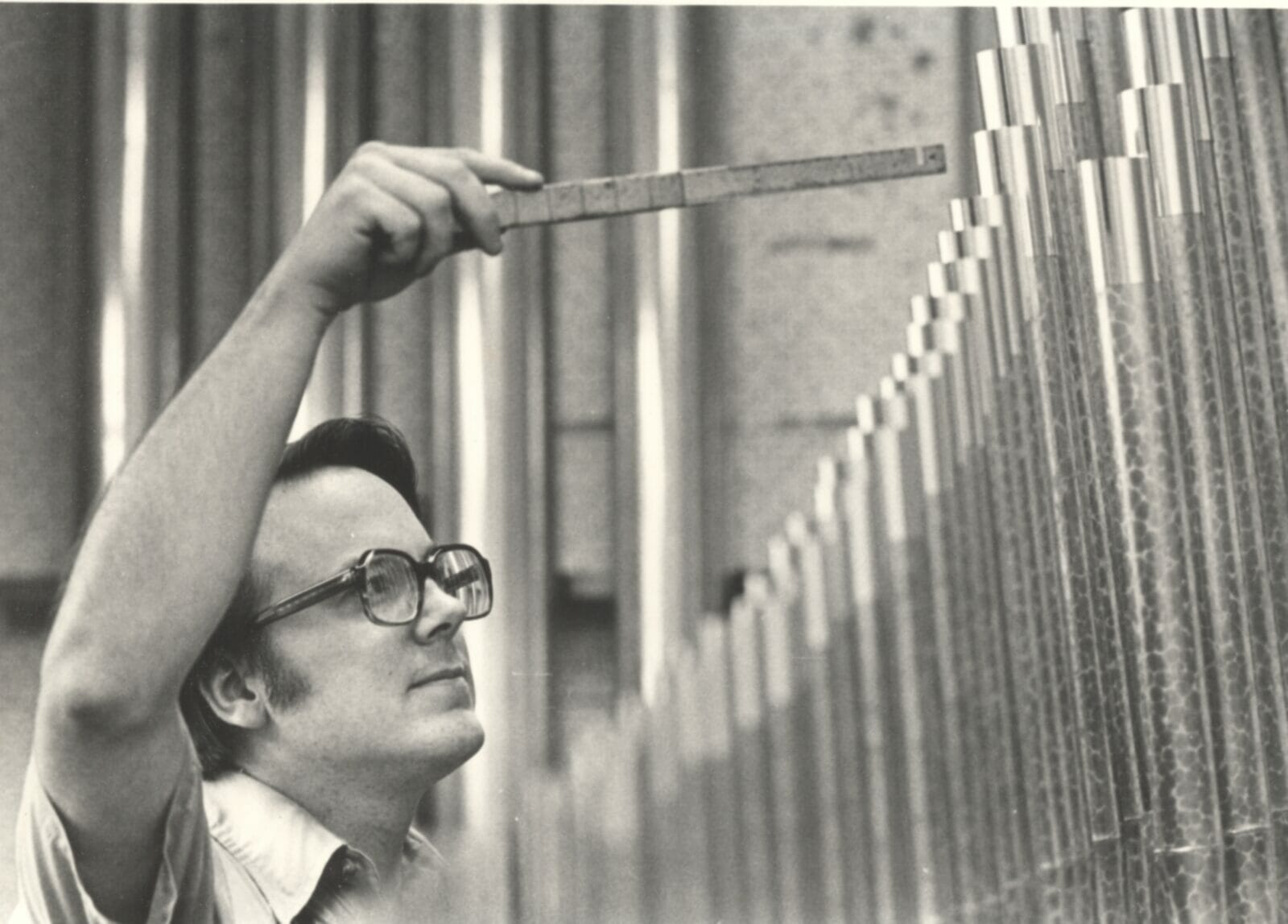 Black and white photo of a man with old, thick glasses holding a tuning instrument up to a row of organ pipes, in front of him.