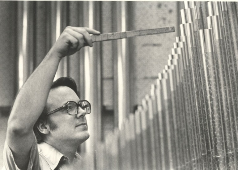 Black and white photo of a man with old, thick glasses holding a tuning instrument up to a row of organ pipes, in front of him.
