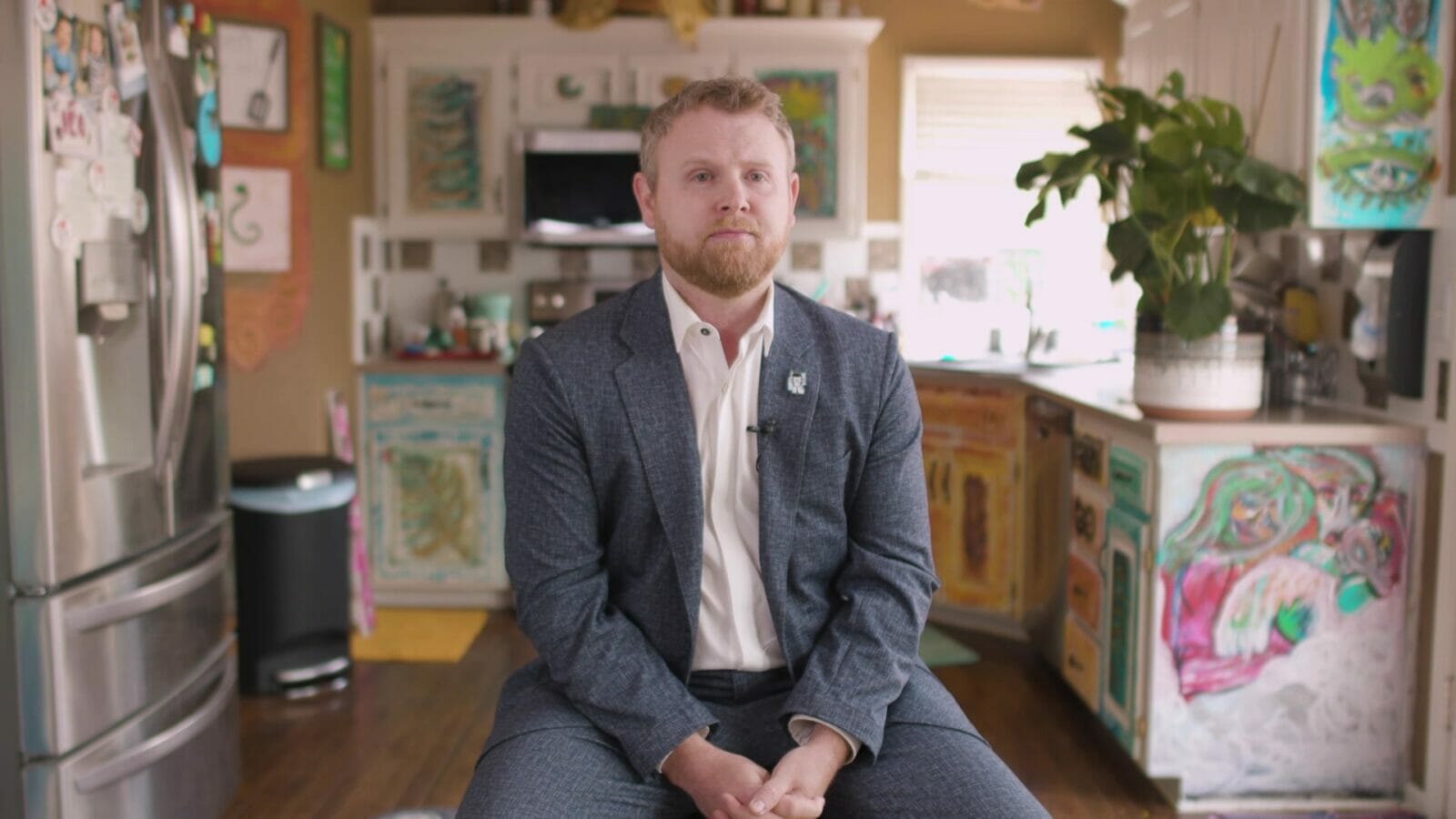 A man in a suit sits in the middle of a colorful kitchen. On his lapel is a pin with the transgender flag.
