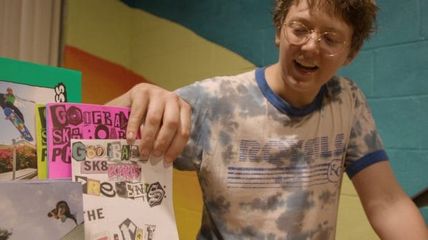 A person in glasses and tie-dyed Royals shirt points to a stack of diy zines.