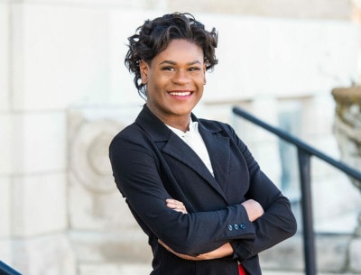 A woman in a blazer and curly hair stands in front of a stone building. She has her arms crossed and smiles at the camera.