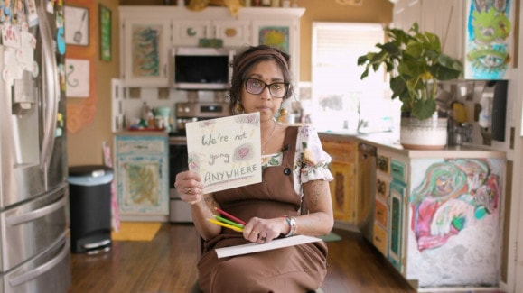 A woman in brown overalls sits in the middle of a colorful kitchen. She holds up a drawing that reads "We're not going anywhere"