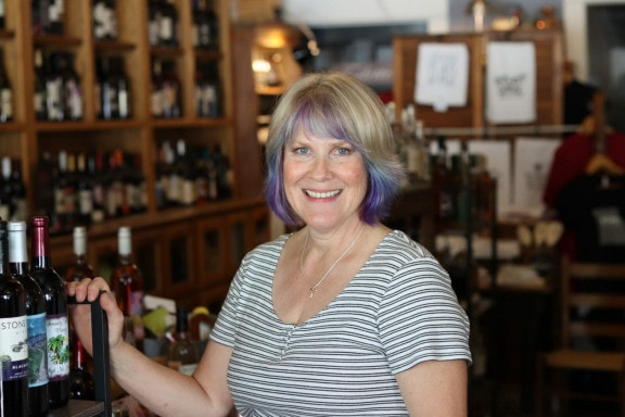 A woman with short hair stands next to shelf with wine bottles, behind her are out of focus shelves with other alcohol bottles.