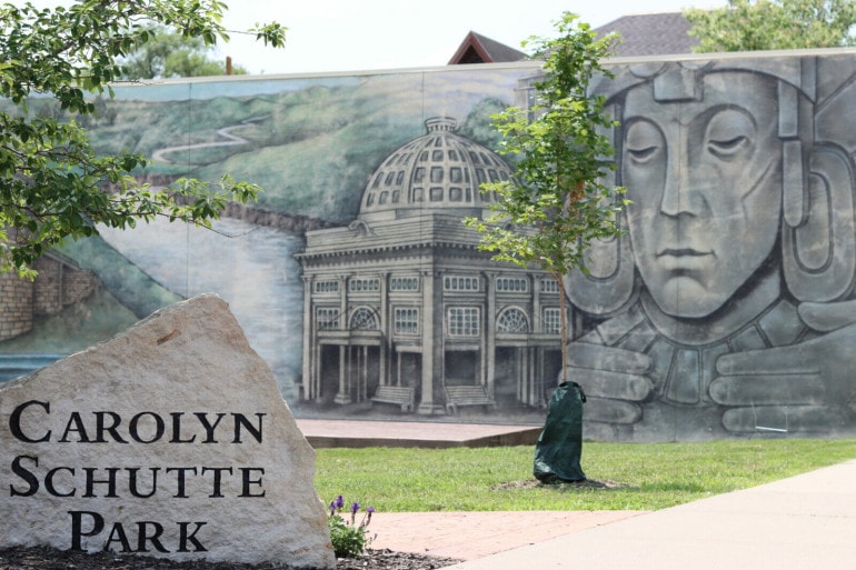 A mural on a wall shows a mayan statue and a stone pergola. A rock in front of the small park reads "Carolyn Schutte Park"