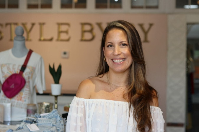 A woman with long hair stands in front of a wall that reads "Style by Ry" next to her is a mannequin styled with a cross body bag and tshirt.
